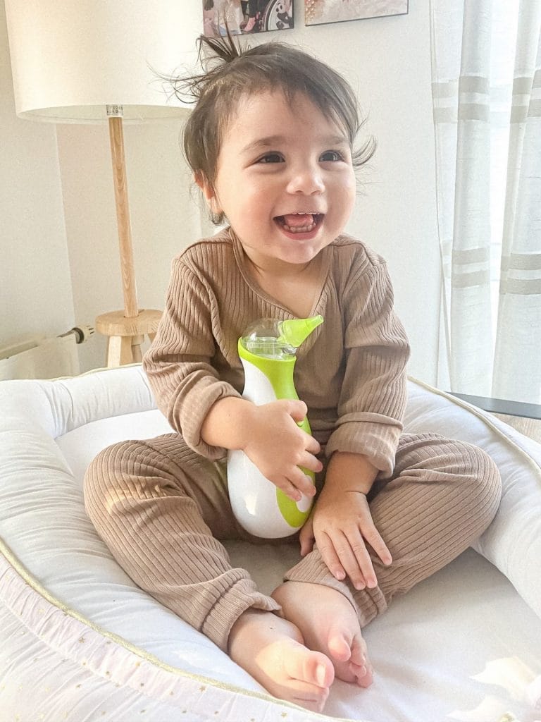 Bambina sorridente in un nido per bambini con un aspiratore nasale elettrico portatile Nosiboo Go in mano