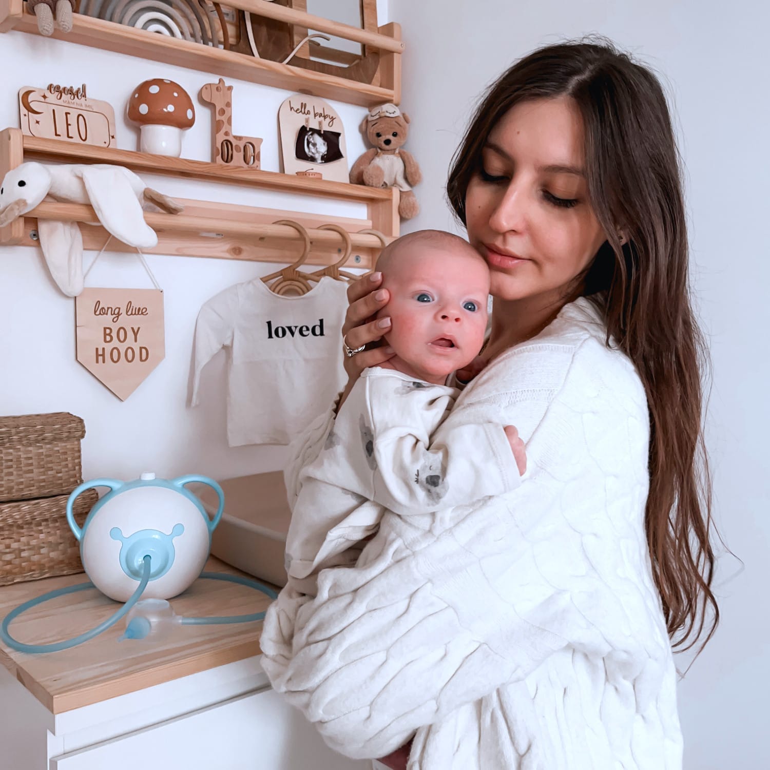 Una bella madre che tiene in braccio il suo bel neonato, in piedi in una stanza per bambini accanto a un aspiratore nasale elettrico Nosiboo Pro.